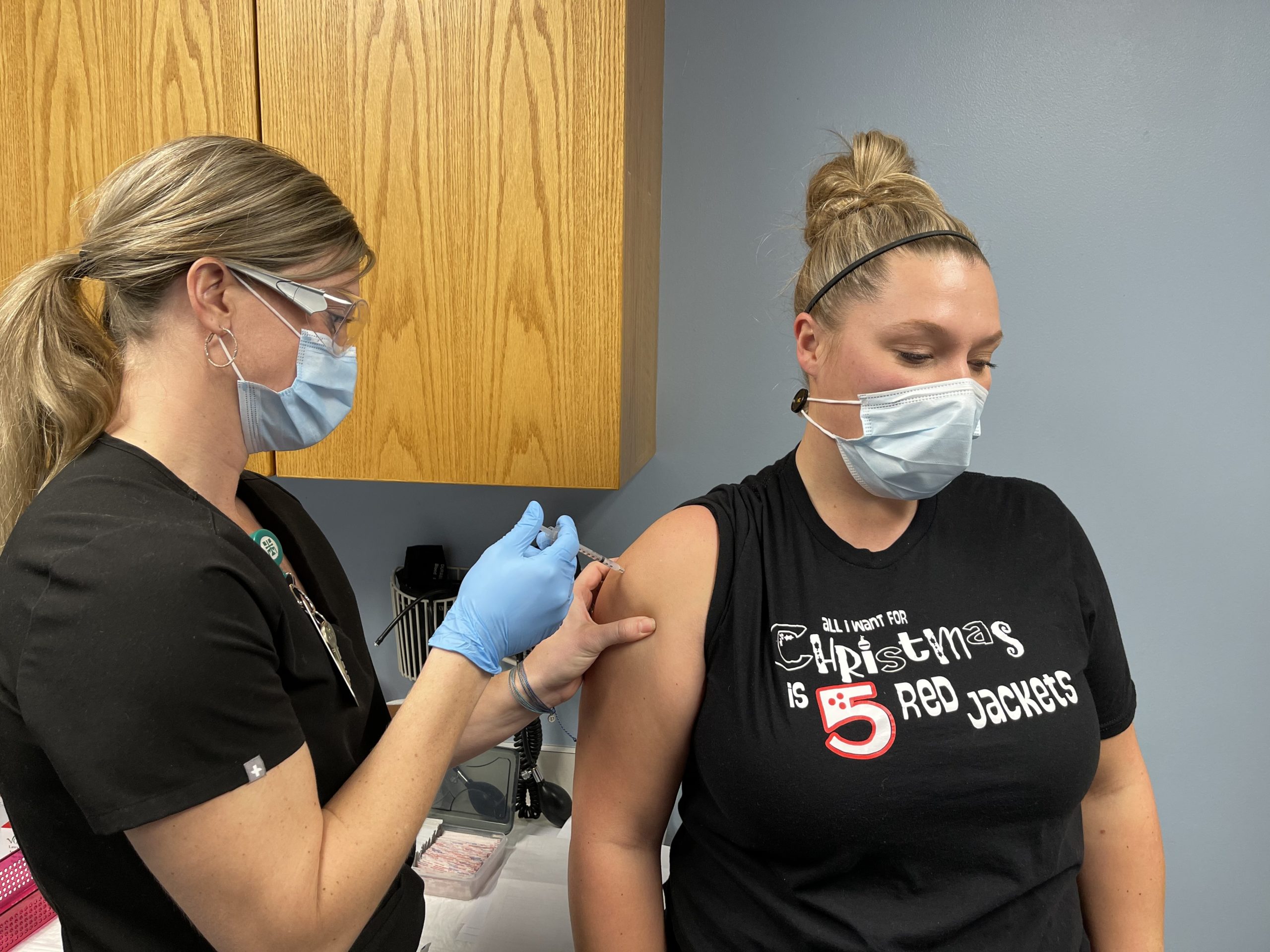 Nurse wearing black scrubs and face mask and goggles giving a COVID-19 vaccine to a woman with blonde hair, wearing a black tee shirt and mask and her hair up in a bun